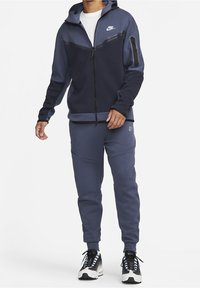 Mörkblå Nike-träningsdräkt med en zip-up hoodie som har en svart bröstpanel, matchande sweatpants med texturerade detaljer och svarta sneakers.
