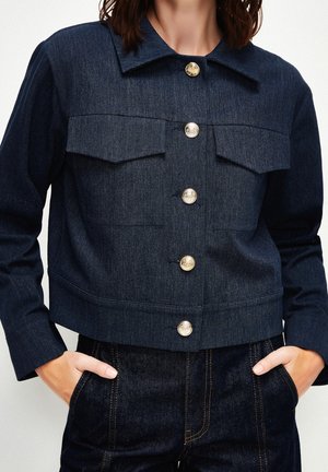 Femme portant une veste en jean bleu foncé avec des boutons dorés et un jean assorti, mains dans les poches avant, fond blanc.