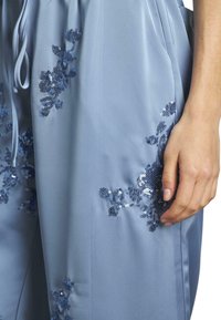 Lichtblauwe satijnen broek met bloemmotief borduurwerk in donkerblauw, versierd met pailletten. Met een trekkoord in de taille en een zachte textuur met een gladde afwerking.