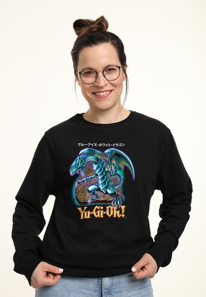 Smilende kvinde med briller iført en sort Yu-Gi-Oh! sweatshirt med en blå drage og japansk tekst.