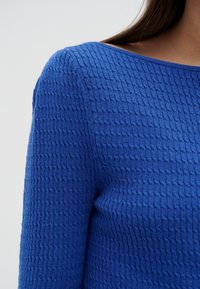 Acercamiento de un suéter de punto texturizado azul en una persona con cabello largo castaño, mostrando detalles de los hombros y el escote.