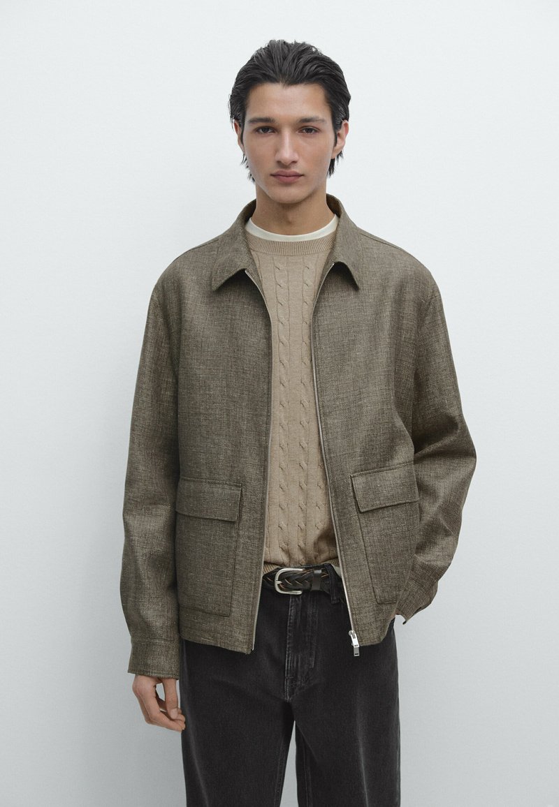 Ung mand med mørkt hår iført en beige kabelstrikket sweater, brun lynlåsjakke, sorte jeans og et flettet læderbælte.