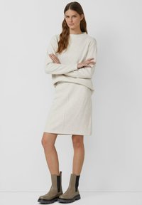 Set van crèmekleurige trui en rok, beide van zachte, gebreide stof. De rok heeft een rechte snit met een zijsplit. Bruine enkellaarsjes met zwarte accenten.