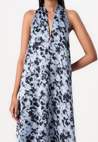 Robe sans manches à col licou avec un décolleté en V profond en tissu bleu clair présentant un imprimé floral abstrait noir, portée par une personne ayant une main sur la hanche.