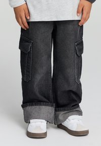 Pantaloni cargo in denim neri con tasche laterali, vestibilità ampia e orli grigi chiari ripiegati. Indossati con sneaker bianche con dettagli grigi.