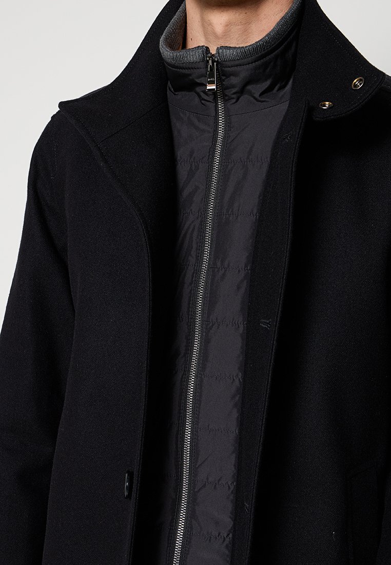 Manteau noir avec un col structuré, comprenant une couche intérieure zippée, des textures de tissu contrastées et des accents en métal argenté.