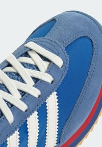 Modrá sportovní obuv s kombinací semišových a síťovaných materiálů, bílými zubatými pruhy a texturovanou podrážkou s červenými a gumovými akcenty.