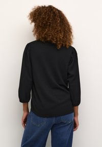 Blouse noire à manches bouffantes trois-quarts, en tissu texturé avec un col rond. Associée à un jean bleu clair.