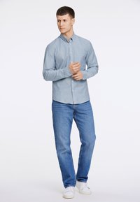 Camisa de botão de manga longa azul claro com colarinho, combinada com jeans azul médio e ténis brancos. Design simples, sem padrões visíveis.