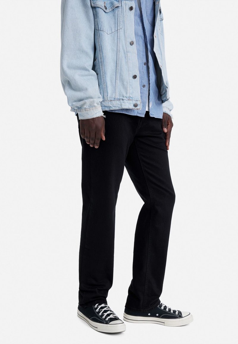 Homme debout portant une veste en jean bleu clair, une chemise en jean bleu foncé, un pantalon noir et des baskets basses noires avec des semelles blanches.