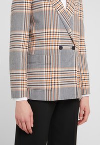 Dubbelzijdige blazer in grijs met een oranje en zwarte ruitpatroon. Bevat twee voorkruipezakken en knopen, gemaakt van textuurstof.