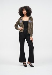 Casaco de veludo verde-oliva com fecho de correr e bordado com o texto "Jeans", combinado com uma blusa preta justa, calças pretas evasê e sapatos pretos de salto.