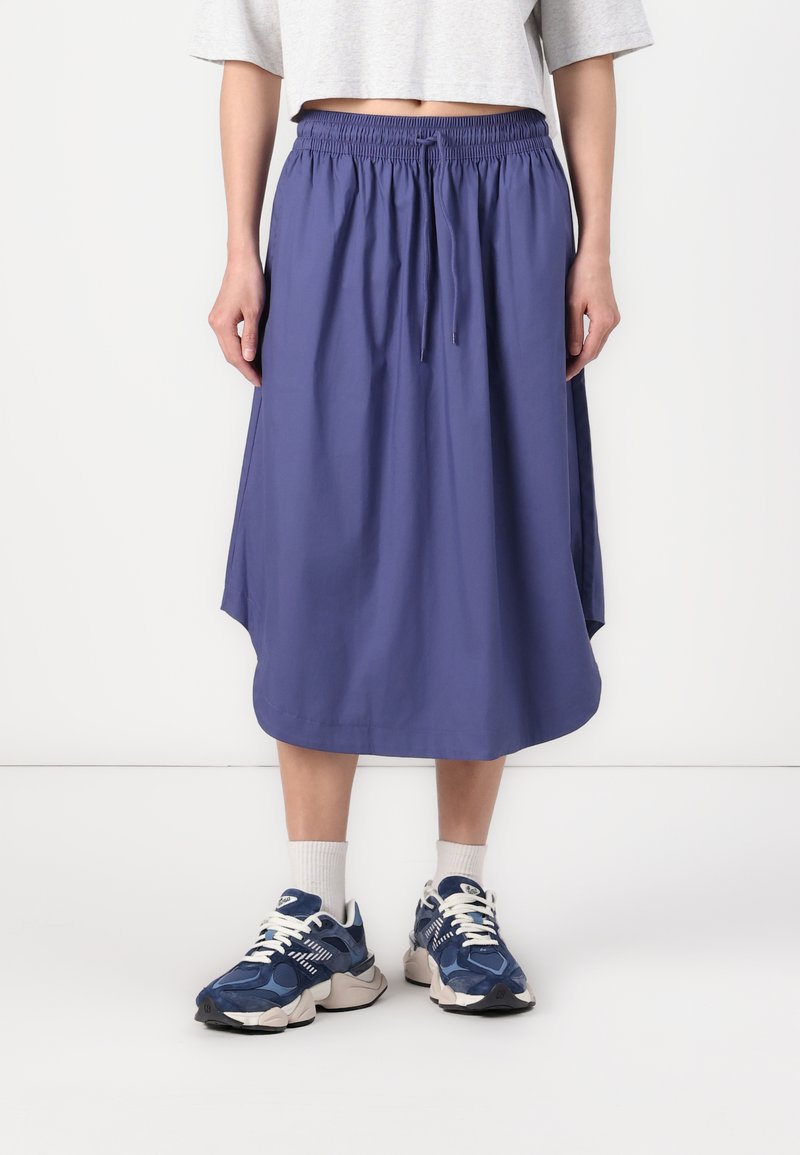 Lilla, knelange skjørt med elastisk midjebånd og snor. Glatt tekstur med en avslappet passform, kombinert med marineblå sneakers og hvite sokker.