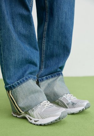 Jambes portant un jean bleu retroussé et des chaussures de sport blanches, debout sur un sol vert sur un fond clair.