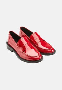 Mocassins en cuir verni rouge avec une pointe arrondie, dotés d'un détail de banderole à l'avant et d'une semelle en caoutchouc noir. Texture lisse et finition brillante.