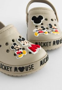 Beigen klomper med Mickey Mouse-tema dekorationer, tekstureret overflade, ventilationshuller og sort "MICKEY MOUSE" tekst rundt om sålen.