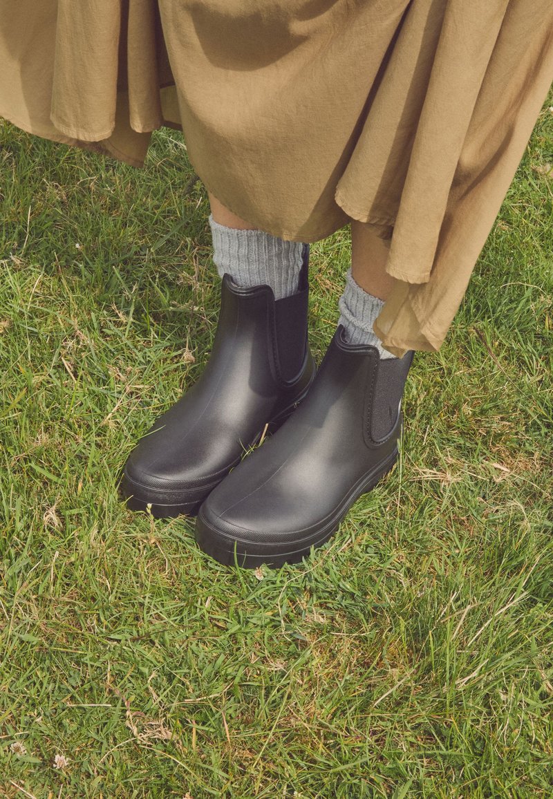 Person wearing black ankle rain boots and gray socks standing on green grass, with a long beige skirt visible above the boots.