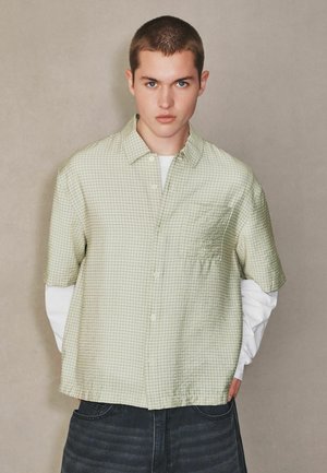 Fiatal személy rövid barna hajjal, világoszöld kockás rövid ujjú inget visel fehér hosszú ujjú ing felett és sötét nadrágot.