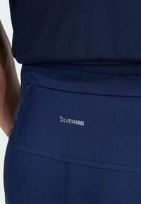 Gros plan sur des leggings de sport bleu marine avec le logo Climacool et une poche zippée sur le côté, portés avec un haut assorti.
