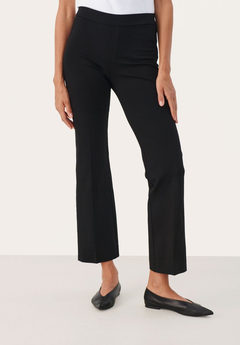 Zwarte, fitted broek met een brede tailleband en een uitlopende pijp. Gemaakt van een gladde, rekbare stof. Draagt met zwarte ballerina's. Minimaal zichtbare naden.