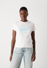 Hvit bomull t-skjorte med blå trykt tekst "Authentic Original Tommy New York City," med korte ermer og klassisk rund hals.
