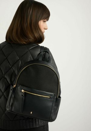 Kobieta w czarnej pikowanej kurtce, nosząca czarny skórzany plecak z złotym zamkiem, pokazana z boku na jasnym, jednolitym tle.