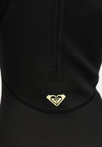 Κοντινή λήψη μαύρου ενδύματος με μαύρο φερμουάρ και μικρό ανοιχτό κίτρινο λογότυπο σε σχήμα καρδιάς με στυλιζαρισμένα πρόσωπα κάτω από τη ραφή του φερμουάρ.