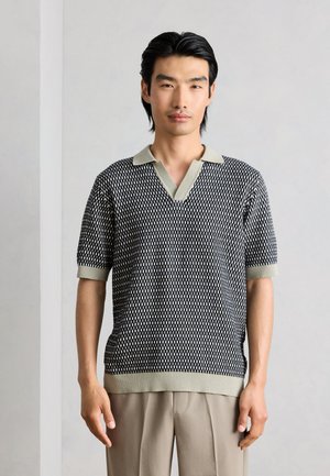 Muž s čiernymi vlasmi, ktorý má na sebe krátky čierno-biely pletený polokošeľu a béžové nohavice, stojí pred jednoduchým pozadím.