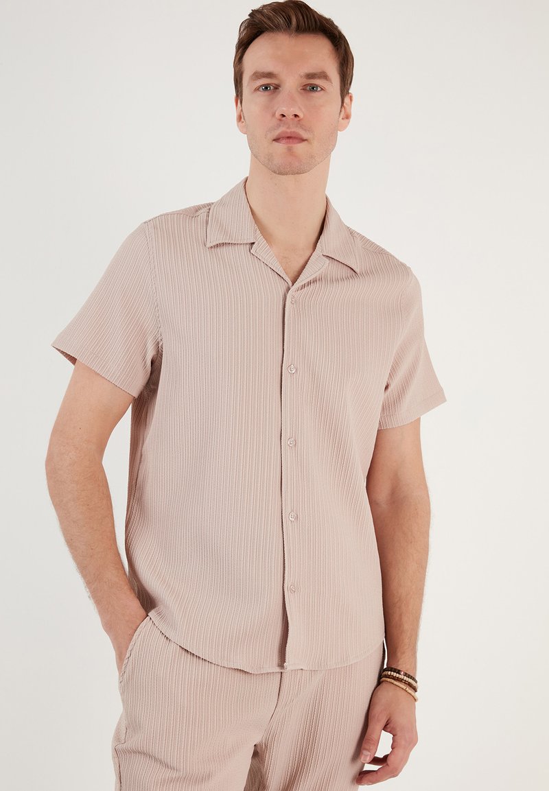 Camisa de manga corta en color rosa claro, confeccionada en tela de canalé. Presenta un cuello y botones en la parte delantera, con un ajuste relajado.