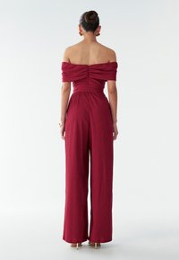 Burgundský overal s odhalenými ramenami s textúrovaným povrchom, priliehavým korzetom a širokými nohavicami, vybavený zadným zipsom a bočnými vreckami.