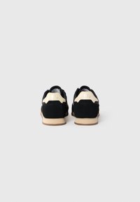 Black suede sneakers with cream accents, rounded shape, rubber soles, and stitched detailing. Features a textured heel with embossed numbers.