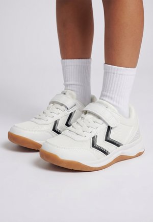 Weiße Sneakers mit schwarzen Chevron-Streifen, Gummisohlen und Klettverschlüssen an Füßen mit weißen, gerippten Socken vor einfarbigem Hintergrund.