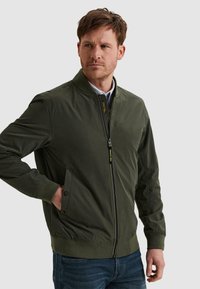 Mann trägt eine dunkelgrüne Bomberjacke mit Reißverschluss und Druckknopftaschen, steht mit einer Hand in der Tasche vor einem schlichten Hintergrund.