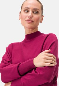 Magenta Strickpullover mit hohem Kragen und langen Ärmeln, ausgestattet mit gerippten Bündchen und einer weichen Textur. Das Model posiert mit verschränkten Armen.