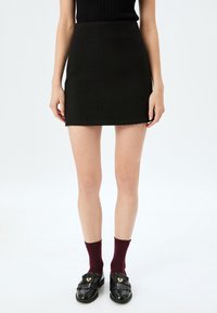 Mini-jupe noire en tissu texturé avec une silhouette ajustée, associée à des mocassins noirs brillants et des chaussettes bordeaux.