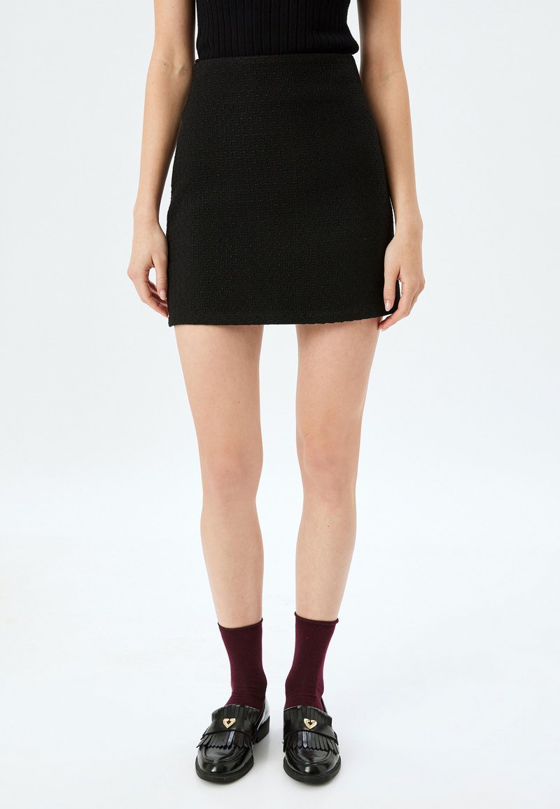 Mini-jupe noire en tissu texturé avec une silhouette ajustée, associée à des mocassins noirs brillants et des chaussettes bordeaux.