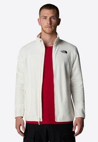 Hombre con una chaqueta de forro polar blanca con cremallera sobre una camiseta roja y pantalones cargo negros, de pie contra un fondo claro y liso.