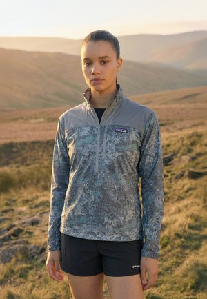 Mladá žena so zviazanými vlasmi, oblečená v vetrovke s maskáčovým vzorom a čiernych šortkách značky Patagonia, stojí na trávnatej, kopcovitej vonkajšej oblasti.