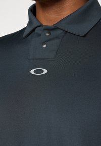 Polo-shirt gris foncé avec un col structuré, deux boutons-pression et un petit logo blanc sur la poitrine gauche. Tissu lisse et léger.