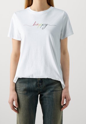 T-shirt blanc à manches courtes avec le mot « happy » écrit en cursive multicolore, porté par une personne debout les mains détendues le long du corps.