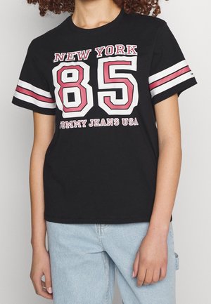 Osoba ubrana w czarny t-shirt z napisem "NEW YORK 85 TOMMY JEANS USA" w wyrazistych różowych i białych literach, w połączeniu z jasnoniebieskimi jeansami.