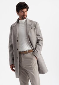 Abrigo de espiguilla gris claro con solapa, botones negros y un bolsillo en el pecho, combinado con un cuello de tortuga blanco y pantalones beige.