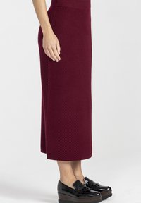 Kobieta w ciemnoczerwonej, prążkowanej spódnicy midi, w połączeniu z błyszczącymi, czarnymi platformowymi loafersami, stoi na tle jasnego, jednolitego tła.