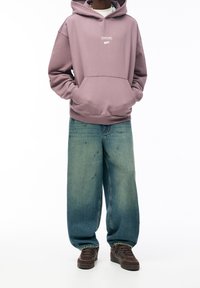 Kapuzensweatshirt in sanftem Lavendel mit weißem Logo, kombiniert mit lockeren, hellblauen Jeans und braunen Sneakers.
