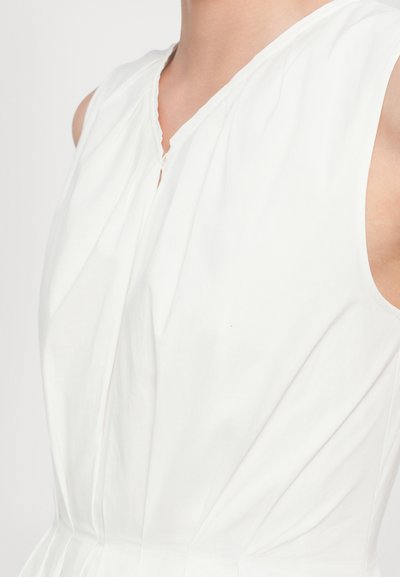 White sleeveless top with a V-neck, featuring pleated detailing and a button closure at the neckline. Smooth, lightweight fabric.