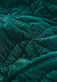 Tkanina pikowana w głębokim zielonym kolorze z miękką, teksturowaną powierzchnią. Zawiera przeszywany wzór, który tworzy wypukły, miękko wyściełany efekt na całej powierzchni.