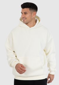 Cremefarbener Hoodie aus weichem Stoff, mit einer Fronttasche, gerippten Manschetten und einer Kordelzugkapuze. Glatte Textur, lässige Passform.