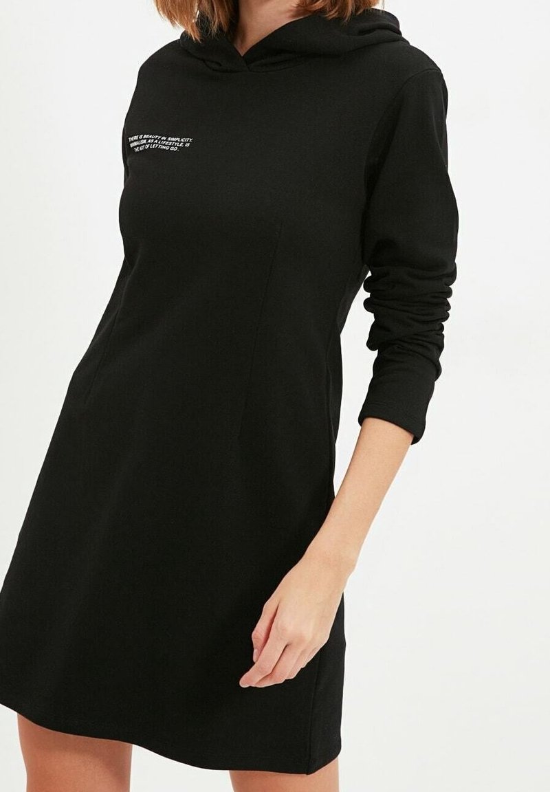Svart hoodieklänning med långa ärmar, tillverkad av mjukt material. Har en tryckt design på framsidan, avslappnad passform och slutar ovanför knäet.