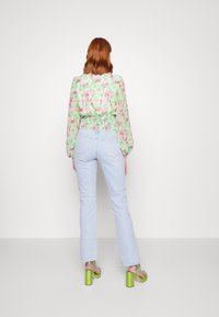 Grüne Bluse mit floralem Muster, langen Ärmeln und smocktem Taillenbund, kombiniert mit hellblauen geraden Jeans und grünen klobigen Fersen-Sandaletten.