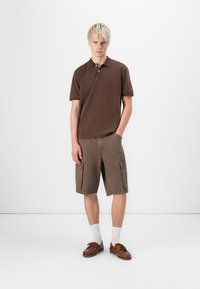 Brown polo shirt with short sleeves, paired with cargo shorts in a darker brown. Model wears brown shoes and white crew socks.